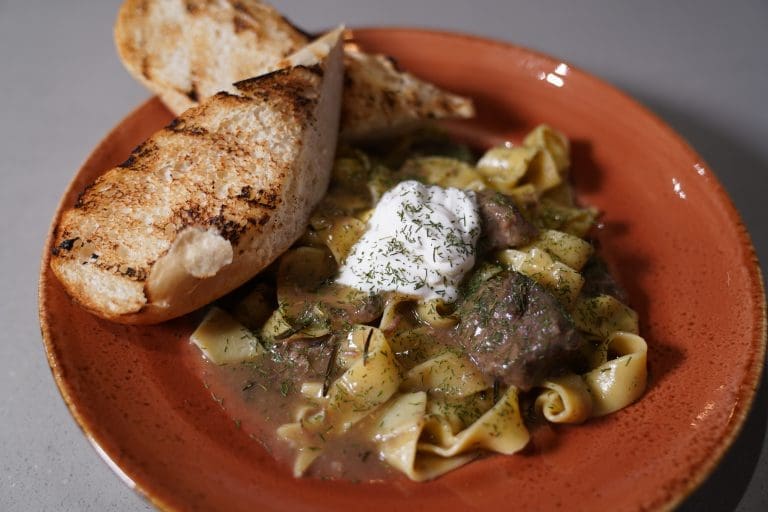 short rib pappardelle with braised short rib gravy pappardelle pasta sour cream parmesan dill and grilled bread in a rust orange bowl on a gray table in a restaurant in sandpoint idaho