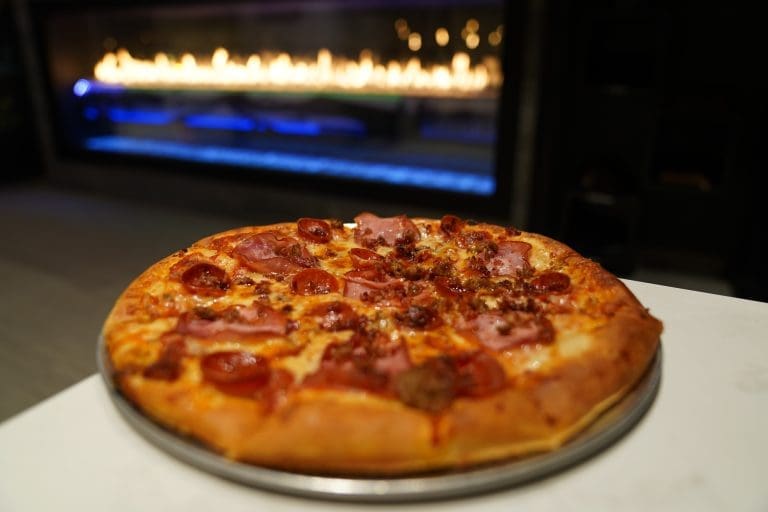 pepperoni bacon crumbled italian sausage and canadian bacon pizza with red sauce and a cheese blend on a gray metal tray on a white counter with a fireplace in the background in savory neighborhood grill in sandpoint idaho