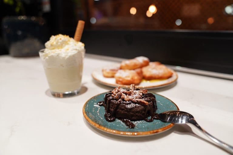 a flourless chocolate torte on a blue plate with a silver spoon laying face down on the tip of the plate a white plate of five powered sugar beignets and a vanilla bean panna cotta on a white counter in savory neighborhood grill in sandpoint idaho