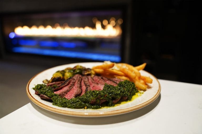 steak frites of grilled 8oz baseball cut steak with chimichurri bleu cheese fries and seasonal vegetables on a white plate on a white counter with a fireplace in the background in sandpoint idaho