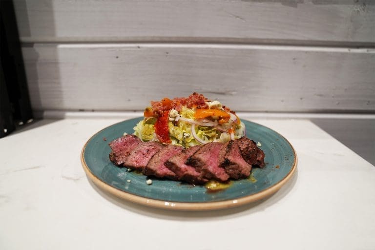 steak wedge salad with iceberg wedge bleu cheese crumbles bacon bites oven roasted and marinated tomatoes grilled sirloin steak and golden balsamic dressing on a blue plate on a white counter in a restaurant in sandpoint idaho